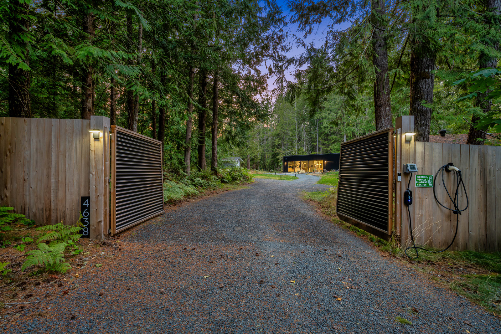The gated entrance at twilight — gravel drive through forest, lanterns glowing, the home visible in the distance